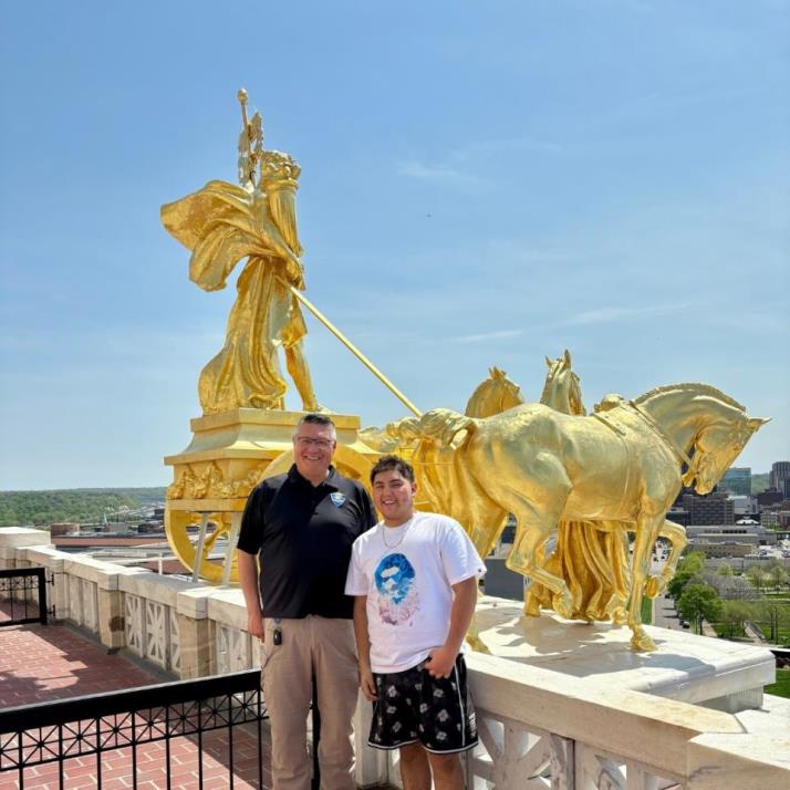 This photo shows two men on top of a building in front of a gold statue of a horse